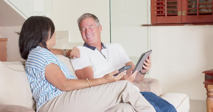 Senior diverse couple relaxing on sofa, using smartphone and tablet at home