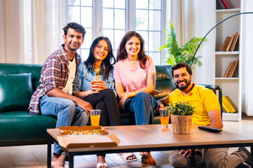 Eating pizza and smiling together, close Indian friends pose for camera in cozy living room