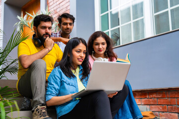 Indian classmates using laptop and books while studying together in friendly outdoor campus setting