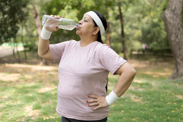 Indian overweight woman in sportswear drinking water after workout at park. Healthy lifestyle,...