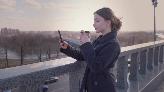 Young teenage girl pausing while walking around town, drinking coffee and taking selfie on smartphone while standing on a bridge overlooking a picturesque cityscape and river