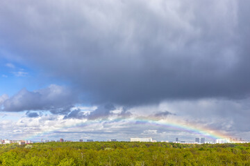 grey rainy clouds and rainbow over green city park
