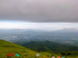 Golden sunrise above green hills, tents scattered on the slope, and a vast valley below the clouds