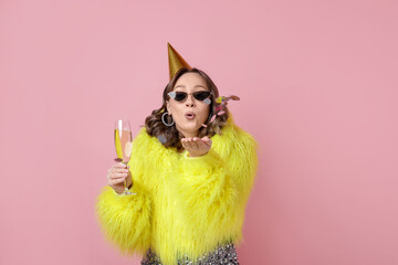 Birthday celebration. Woman with glass of sparkling wine blowing confetti on pink background