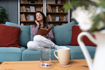 Happy young woman relaxing on comfortable sofa reading interesting book and listening to music with...