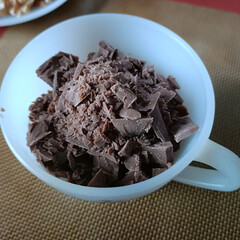 A white mug brims with coarsely chopped milk chocolate on a brown surface. Rich chunks and fine shavings catch natural light, hinting at delicious baking in progress.