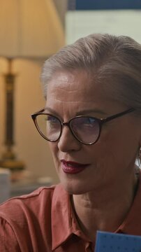 Vertical medium close up of mature female author in glasses speaking deeply during intimate bookstore gathering, surrounded by warm lights and stacked novels