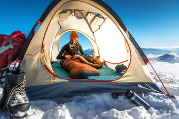 Young woman inside a tent on a snowy mountain, adjusting her sleeping bag on a sunny winter morning. A backpack and mountaineering boots are visible outside the tent © XArt