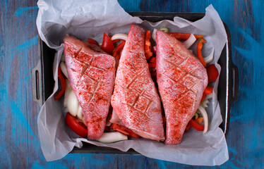 Raw sea bass or pike perch with crosswise cuts on vegetables in a baking tray on a blue table....