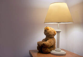 Teddy bear looking at a lit lamp on the nightstand. Photograph with copy space.
