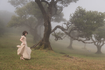 woman dancing in white dress on bare feet on grass under laural trees of Fanal Forest, Madeira, in morning mist