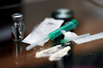 Syringes filled with liquids and powdered substances are placed on a reflective surface. The environment is dimly lit, suggesting a clandestine activity occurring at night.