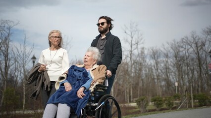 An elderly mother in a wheelchair smiles while her daughter and grandson take her for a joyful walk outside on a beautiful spring day surrounded by nature.