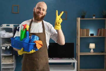 Bearded man janitor showing victory gesture looking at camera holding basket with cleaning supplies for household chores