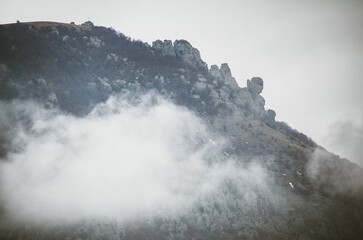 Rugged Crimea mountain peaks shrouded in mist create mystical ambiance. Overcast lighting casts soft gray hues on landscape. Rocks tower prominently, evoking timelessness