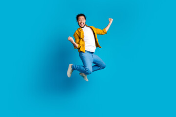 Obraz premium Energetic young man in yellow shirt joyfully jumping against a bright blue background expressing happiness and confidence