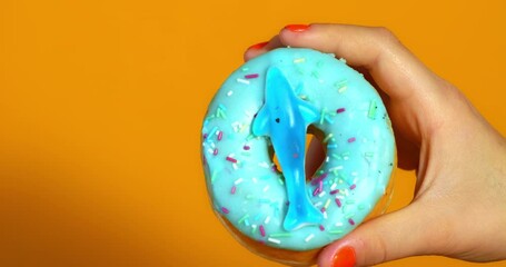 A child’s hand holds a blue frosted donut topped with colorful sprinkles and a gummy shark, set against a vibrant orange background.