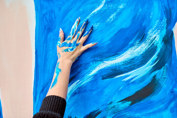 Adult female hand covered in vibrant blue paint creates expressive strokes on large canvas. Bold colors and dynamic movement convey creativity and emotion