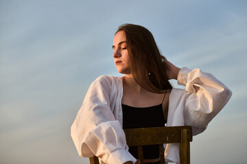 Young woman in white blouse and black top sits on wooden chair by ocean. Golden hour light creates...