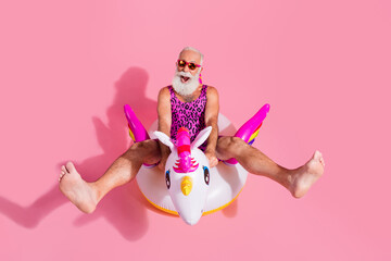Elderly man in funky leopard print swimsuit posing with unicorn float on pink background