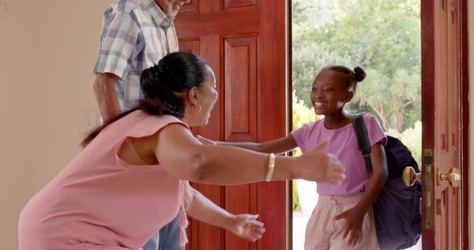 Diverse grandparents welcoming granddaughter with backpack at home, sharing joyful hugs