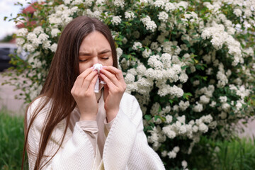 Woman with paper tissue suffering from seasonal pollen allergy near blossoming shrub on spring day,...