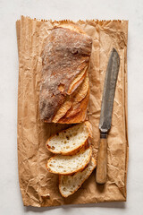 A baguette with thick slices beside it rests on a rustic paper bag. Next to it, a bread knife used for cutting.