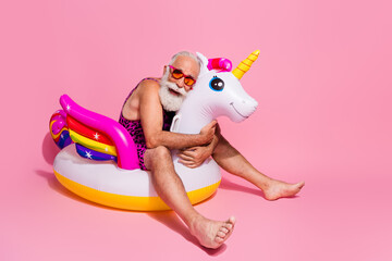 Elderly man in leopard print swimsuit enjoying holiday fun with inflatable unicorn pool float on pink background