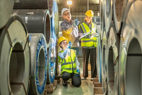 Industrial manufactu factory scene with diverse workers in safety gear inspecting large rolled steel coils and industrial equipment in a modern production facility