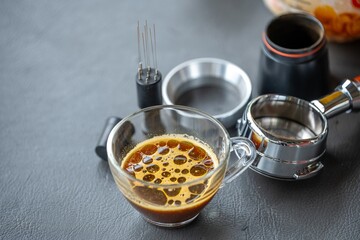 Elegant coffee extraction setup with a glass cup containing brewed coffee topped with foam, a metal pour-over brewer, stainless steel funnel, filters, and a drip kettle on a dark surface