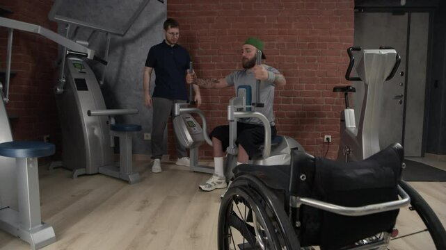 Bulked gym resident with disability having workout for upper limbs under personal trainers control
