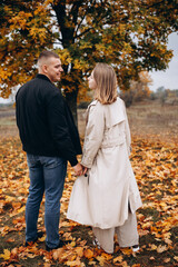 Fototapeta premium Young couple holding hands while walking in an autumn park. Smiling lovers enjoying the moment surrounded by golden leaves.Smiles, warmth, and romance in a beautiful natural fall setting.