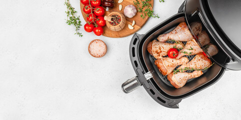 Raw seasoned chicken drumsticks in air fryer with fresh ingredients on white kitchen table. Long...