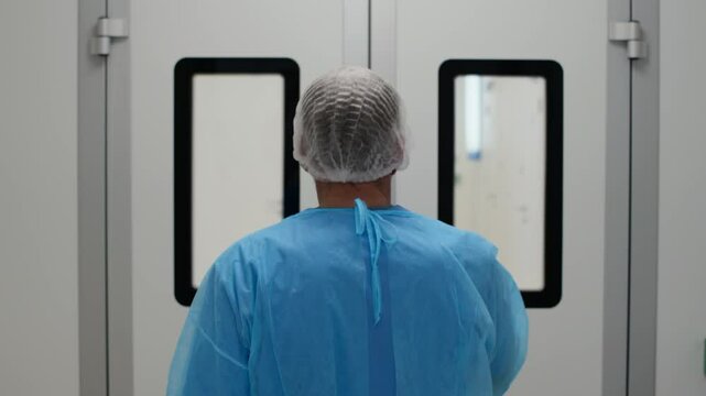 Back-view medium shot of male surgeon in full sterile attire walking down clinical hallway through double doors leading to sterile surgical area