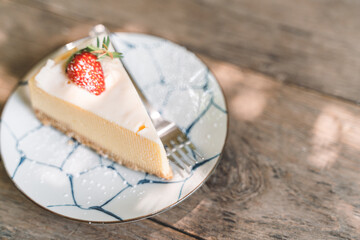 Classic slice of cheesecake with a fresh strawberry on top, served on a patterned ceramic plate with a fork, placed on a rustic wooden table.