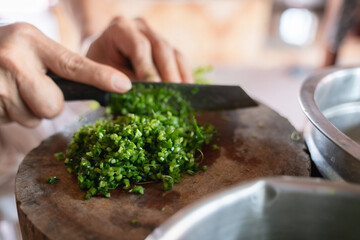 Close-up of hands finely chopping green herbs on a wooden cutting board with a sharp kitchen knife, preparing ingredients for cooking.