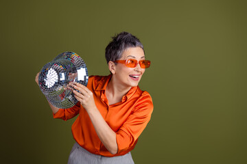 Charming senior woman in orange shirt holding disco ball against green backdrop, embracing joy and...