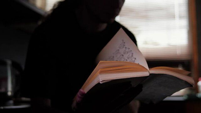 A man, viewed from behind, sits in a dark room, illuminated only by a headlamp he is wearing, which shines light onto an open book he is reading. The scene conveys a sense of quiet concentration