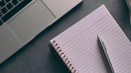 Overhead view of a lined notebook with pen beside laptop on dark desk