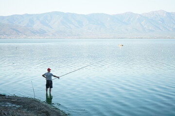 A man is fishing in a calm, light teal-colored lake. The water's surface is slightly rippled. The distant shore line is a light grayish-tan, and low, hazy mountains form a backdrop, also light grayish