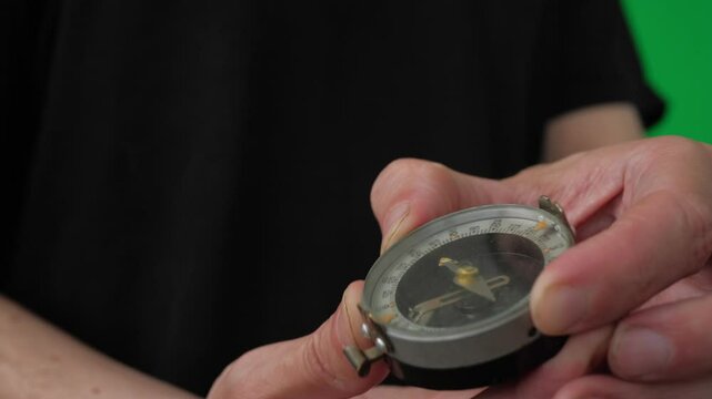 A close-up shot of a hand holding a compass, a vintage military-style compass with a metal casing and a glass cover over the dial. The compass features a sighting mechanism and a thumb loop