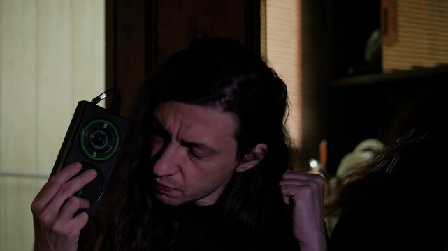 A man with long hair, eyes closed, listens intently to a portable radio, holding it close in a dimly lit room, suggesting deep concentration or a private moment