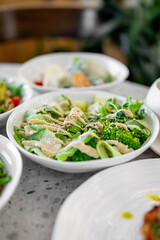 Fresh green salad with lettuce, broccoli, cucumber ribbons, and sesame seeds, drizzled with creamy dressing, served in a white bowl on a stone countertop. Blurred dishes in the background