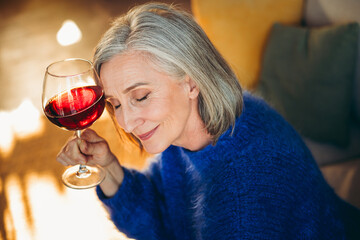 Elderly woman indulging in peaceful relaxation holding a glass of red wine, enjoying sunlight indoors on a cozy autumn day