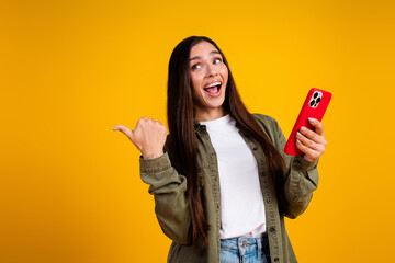 Charming young woman with long brunette hair excitedly holding a smartphone in front of a yellow...