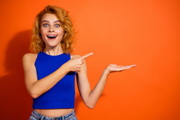Cheerful woman with bright smile points to the side on vibrant orange background wearing casual blue top representing excitement