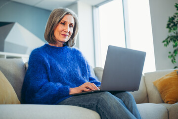 Mature woman relaxing at home on sofa while using a laptop in a cozy living room on a bright sunny day