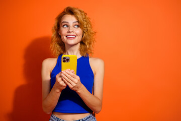 Young red haired woman in blue top holding yellow phone against vibrant orange background,...