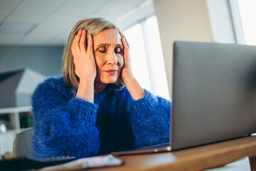 Mature woman in casual sweater indoors experiencing stress at work with her laptop in a brightly lit room