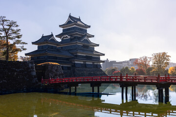 Obraz premium Matsumoto Castle and its red bridge reflected in the still moat water, surrounded by autumn foliage and illuminated by soft morning light through a light haze